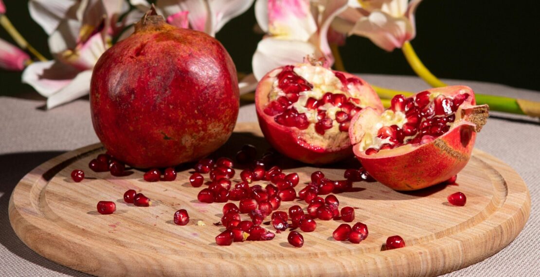 Close-up of whole and sliced pomegranates with flowers on a wooden tray, vibrant and fresh setting.